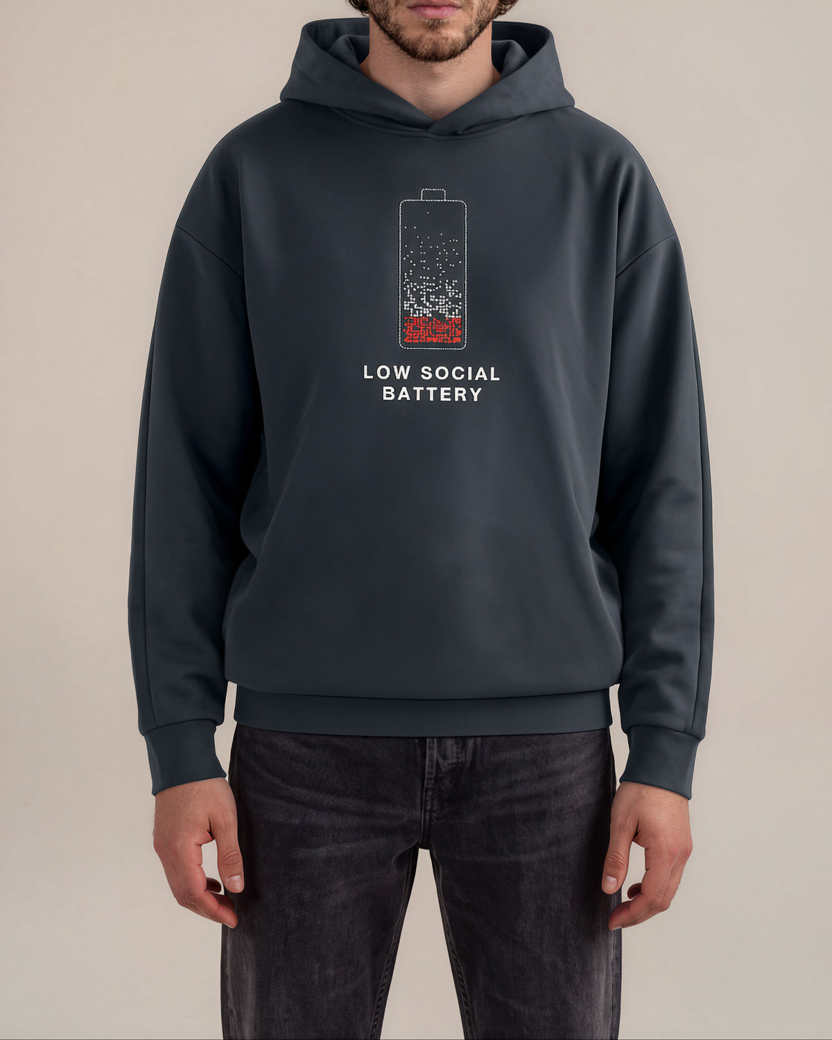 A man wearing a dark slate gray hoodie with a graphic of a battery that is half-filled with red liquid and the text 'LOW SOCIAL BATTERY' printed below it, paired with dark gray jeans against a plain beige background.