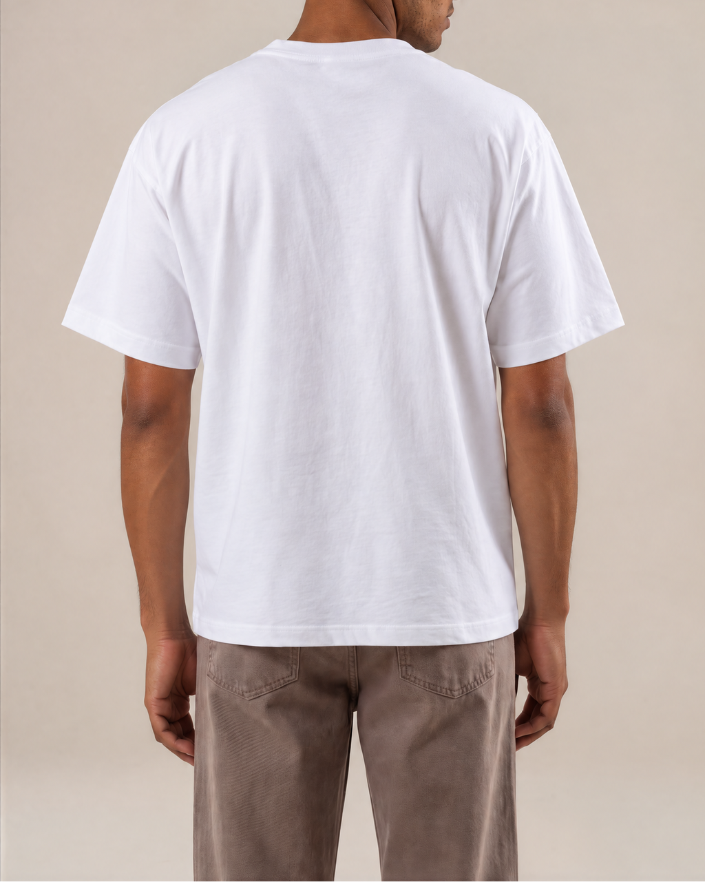 A man viewed from the back wearing a plain white short-sleeve crewneck t-shirt and brown pants, standing against a solid beige studio background.