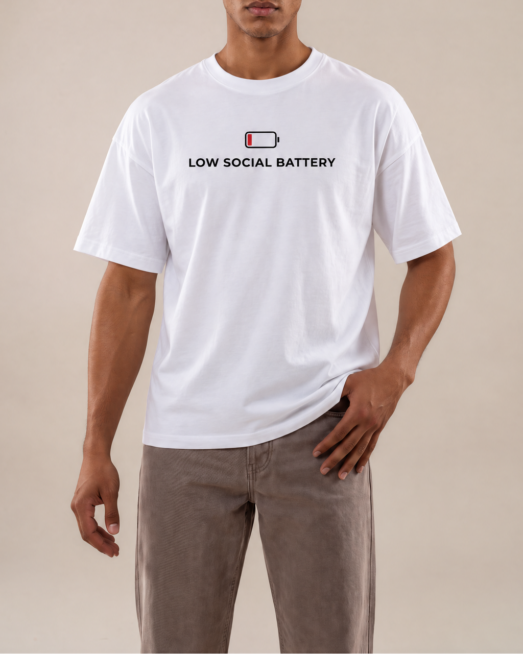 A man modeling a white short-sleeve t-shirt with a black graphic that reads 'LOW SOCIAL BATTERY' and a small battery icon. He is wearing brown pants against a solid beige studio background.