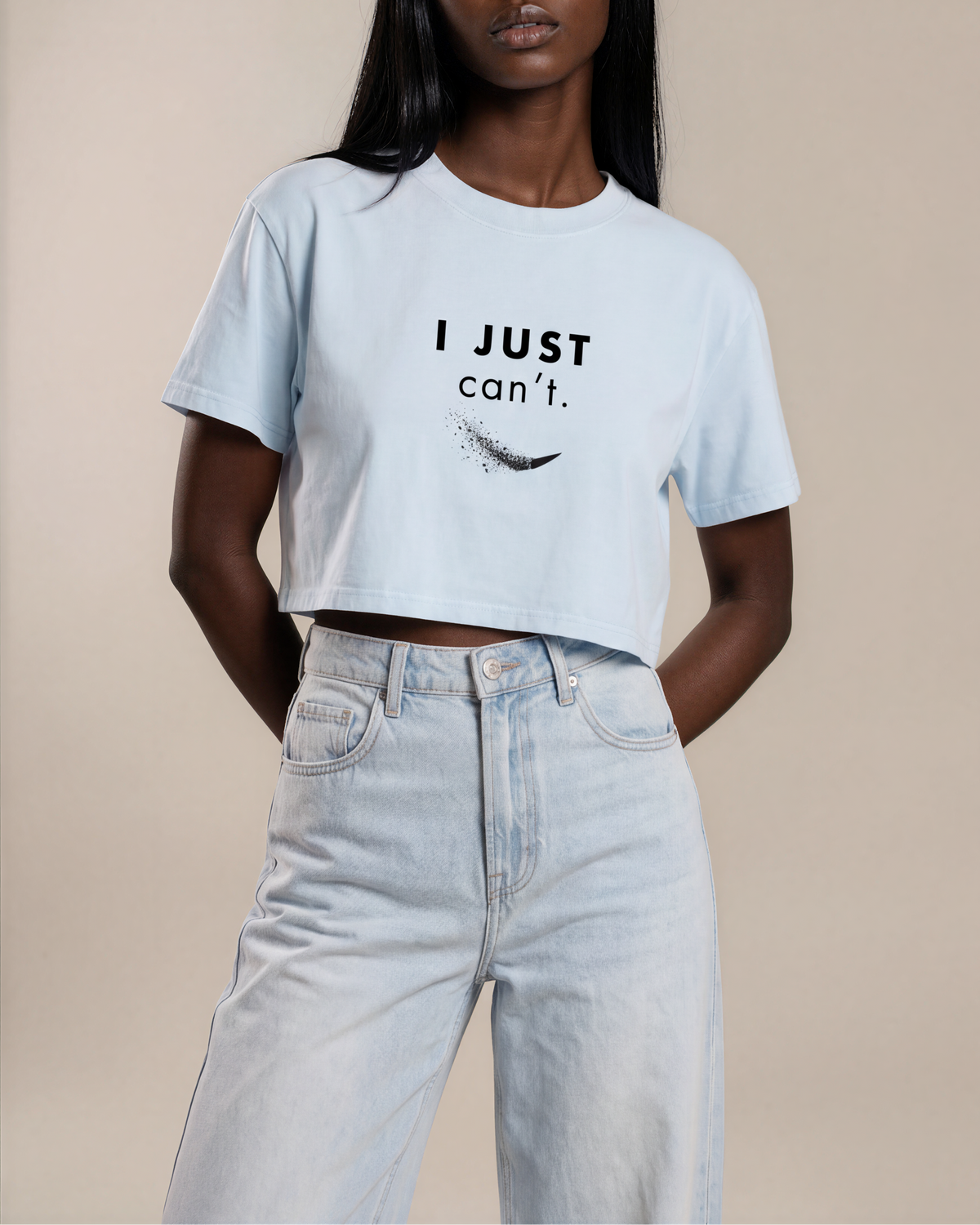 A woman modeling a light blue cropped t-shirt with the text 'I JUST can't.' and a feather graphic printed on the front, paired with light-wash jeans against a solid beige background.