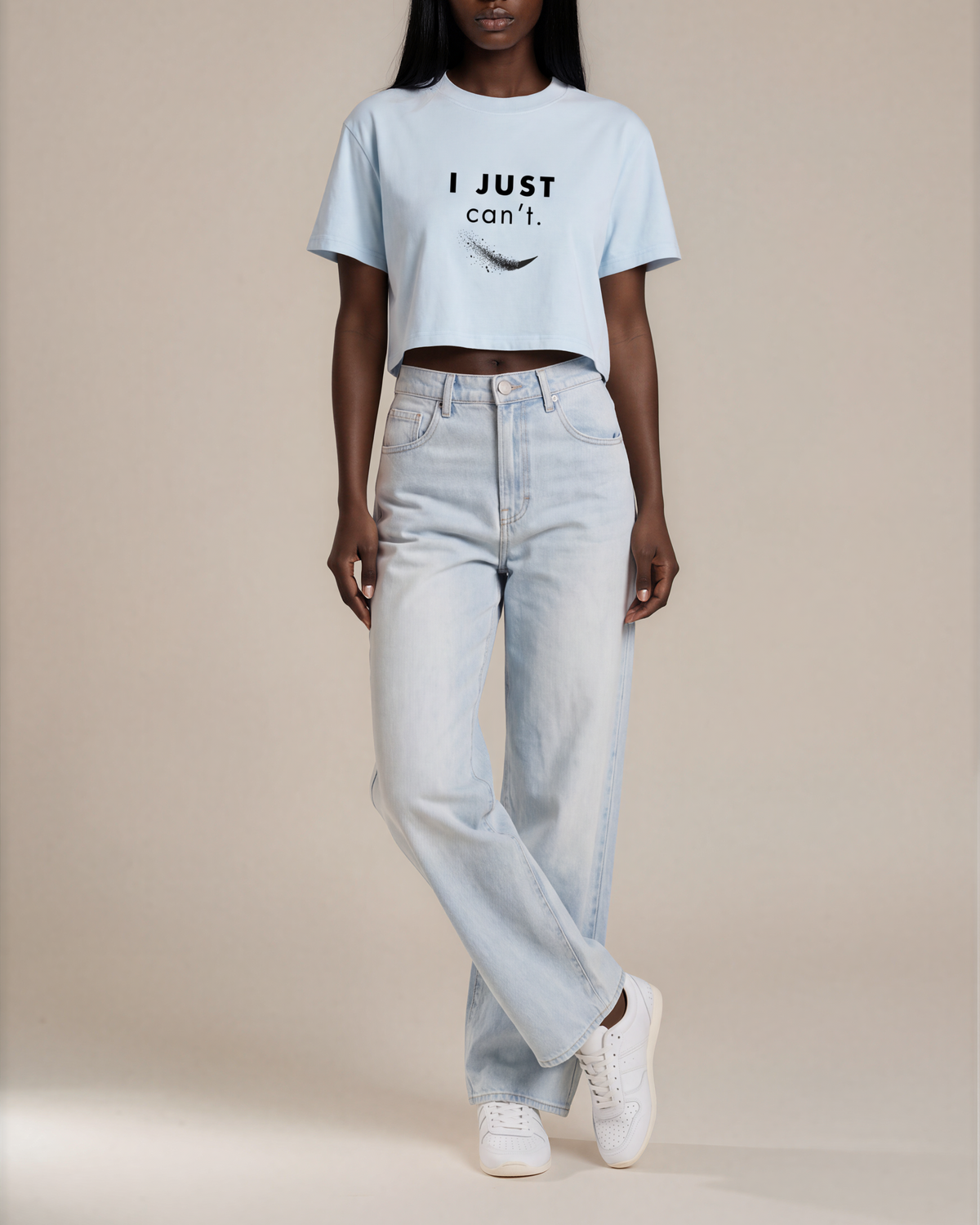 A model stands against a plain beige studio background wearing a light blue cropped T-shirt with the text 'I JUST can't.' printed in black on the chest, light-wash high-rise jeans, and white sneakers.