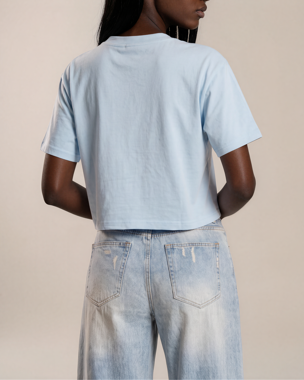 A model shown from the back wearing a light blue cropped t-shirt and light-wash distressed jeans against a solid beige studio background.