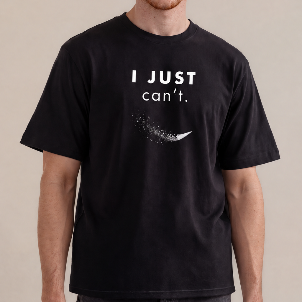 A man wearing a black graphic t-shirt with the text 'I JUST can't.' and a small white graphic below it, paired with dark gray jeans, standing against a neutral beige background.