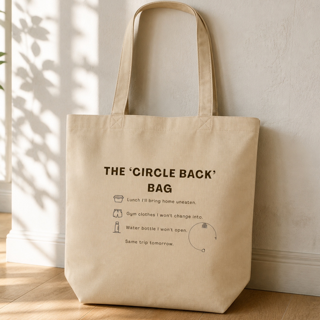 A natural canvas tote bag with printed black text and illustrations reading 'THE 'CIRCLE BACK' BAG' and small icons, leaning against a white wall on a light wood floor in a sunlit room.