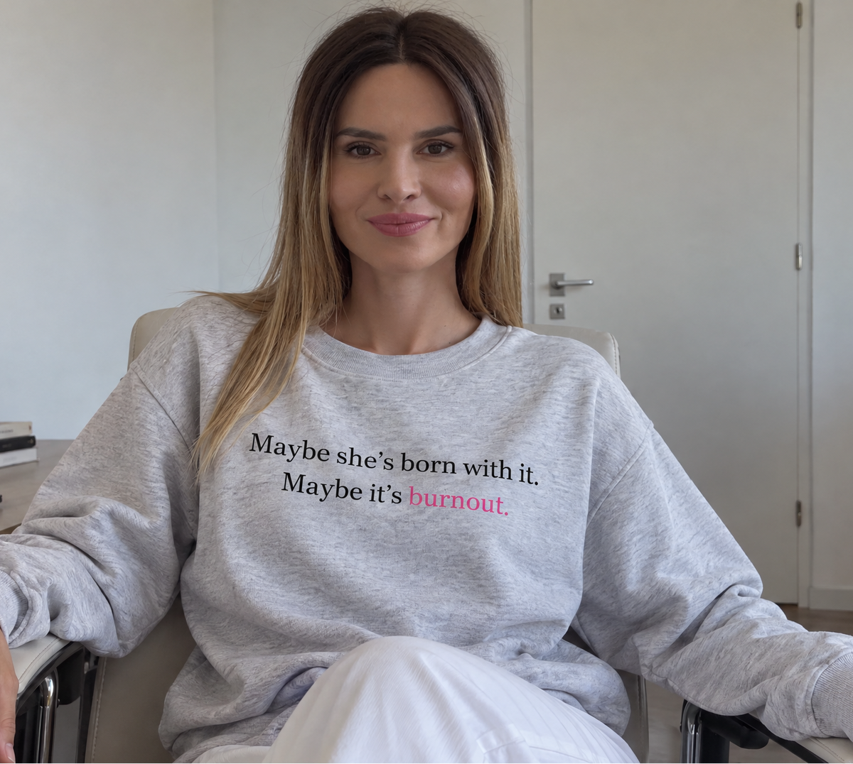 A woman sitting in a chair wearing a light gray crewneck sweatshirt with the text 'Maybe she's born with it. Maybe it's burnout.