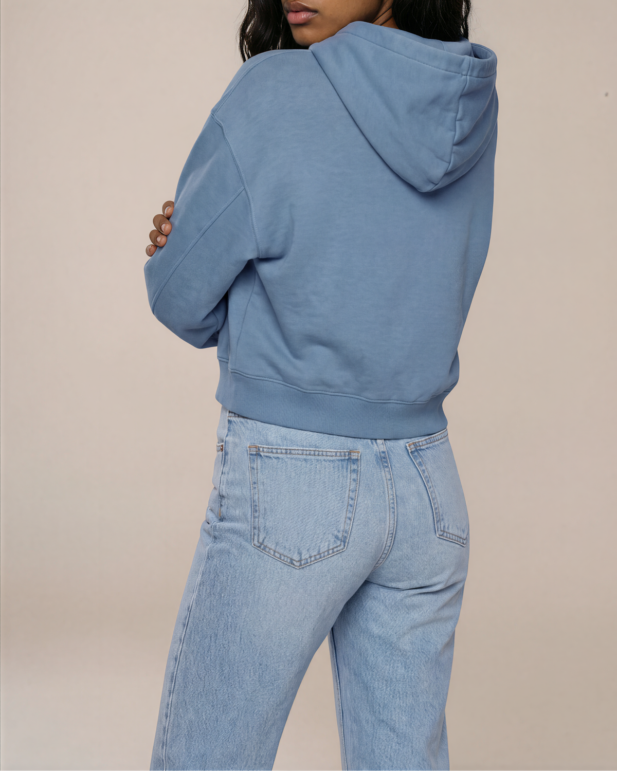 A woman wearing a dusty blue cropped hoodie and light-wash jeans, photographed from the back against a solid beige studio background.