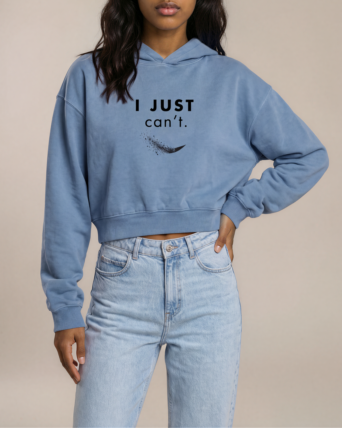 A woman modeling a light blue cropped hoodie with the text 'I JUST can't.' and a feather graphic printed on the chest, paired with light-wash jeans against a solid beige studio background.